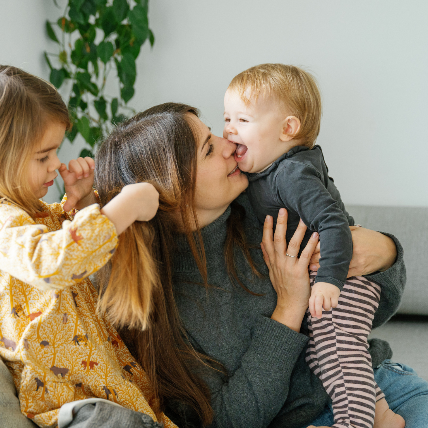 Woman sitting on a couch with two young children, one child is kissing the woman's cheek.