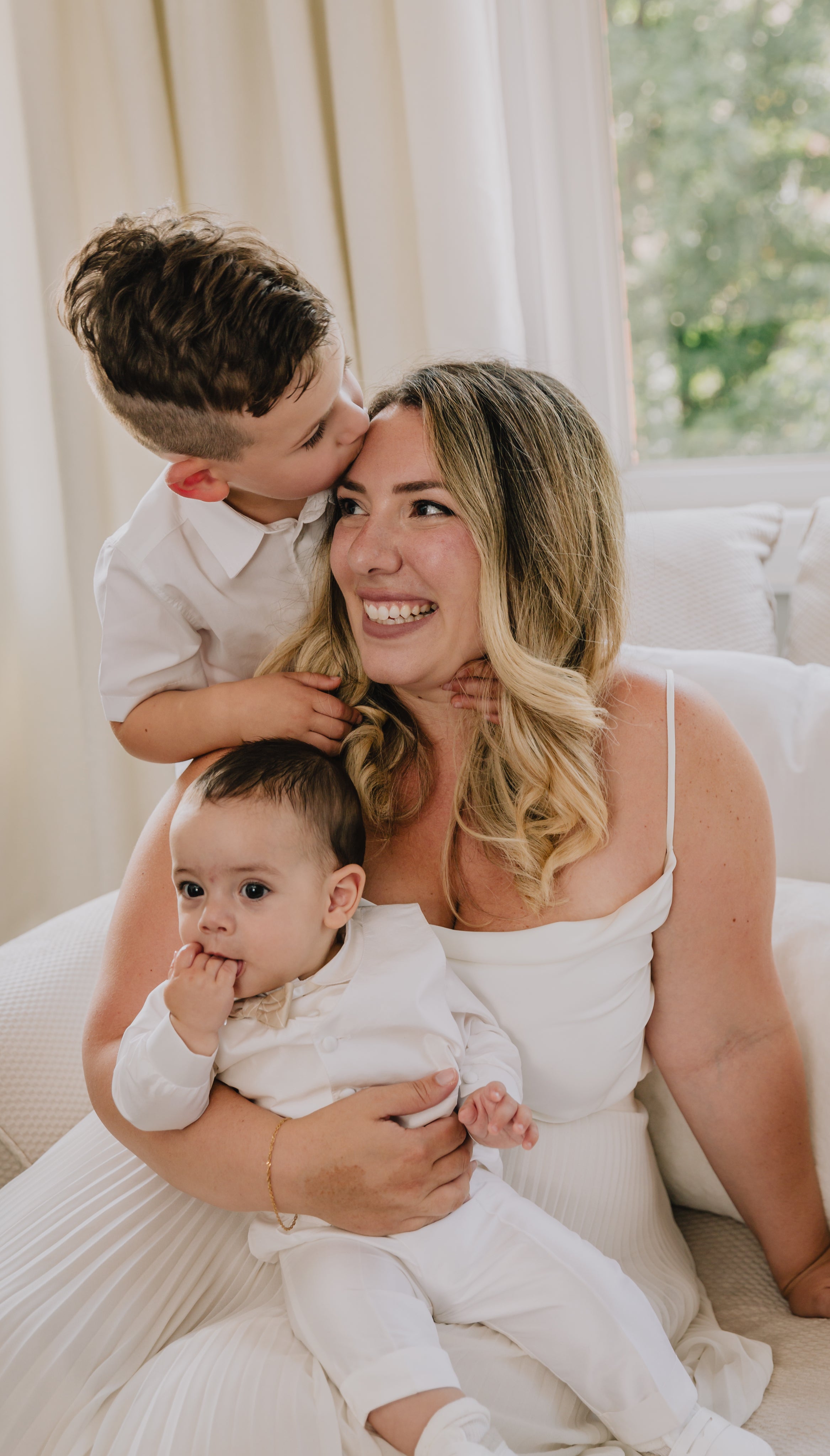 Mum sitting on white sofa holding baby and second child kissing her forehead